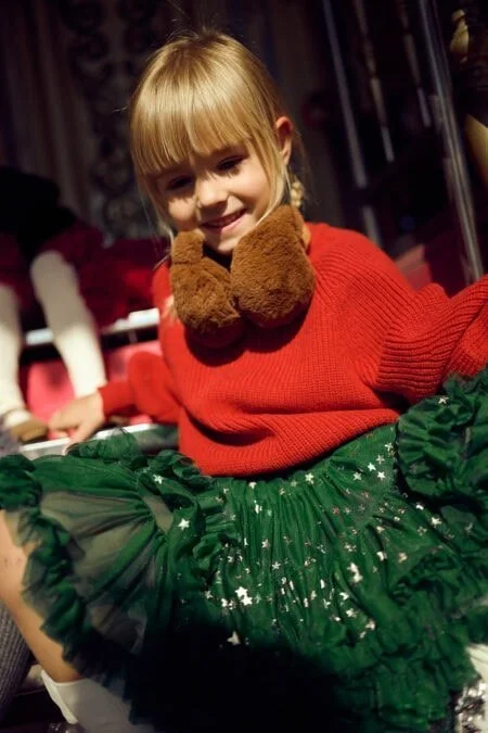 Niña con suéter rojo y falda de tul verde con estrellas doradas, usando un cuello de peluche marrón.