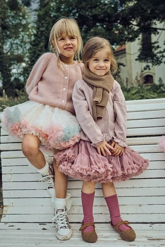 Dos niñas sonríen en un banco del parque. Llevan un jersey de punto rosa y una falda de tul blanca.