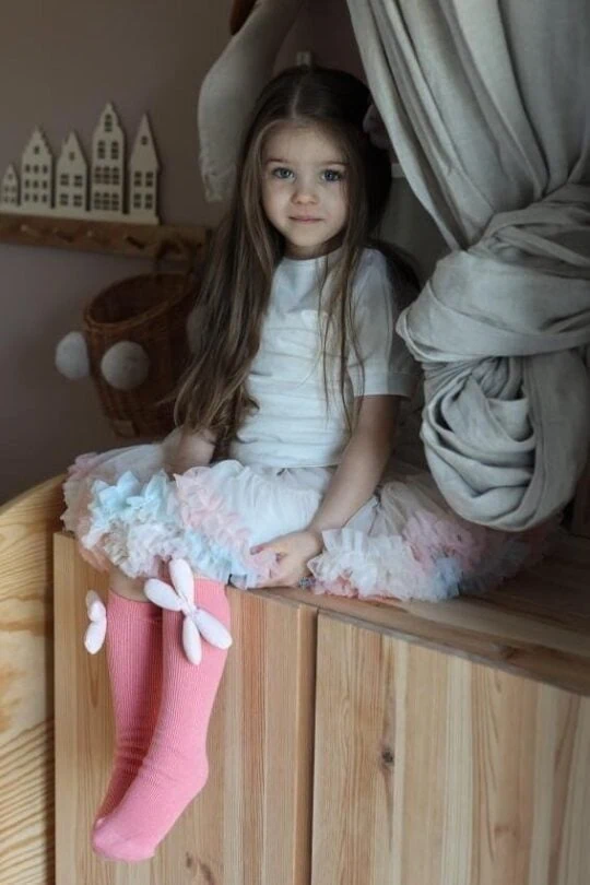 Girl in pink knee socks and colorful tulle skirt sits in room