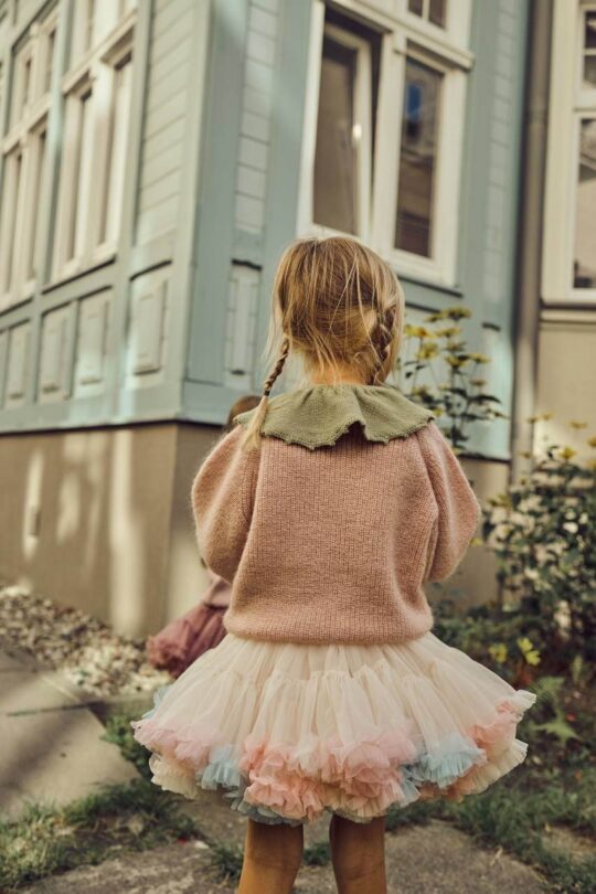 Niña de espaldas con cuello decorativo de lana, jersey de punto rosa y falda de tul blanco